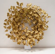 a wreath made out of gold leafy branches on a white pedestal with a white wall in the background