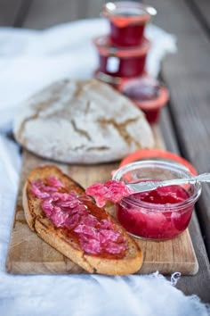 a wooden cutting board topped with bread covered in raspberry jelly next to two jars of jam