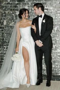 a bride and groom standing next to each other in front of a brick wall wearing tuxedos