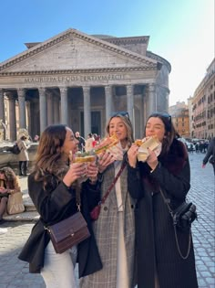 three women are eating sandwiches in front of a building with columns on the side walk