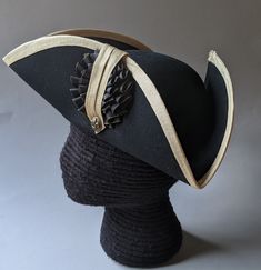 "This military style three cornered tricorn hat is made from top quality wool felt. The edges have been trimmed even and the sides held up or \"cocked\". It has been trimmed around the brim with gold fold-over braid. It is also available with black or white braid. Plain untrimmed hats are listed separately. Gold tone of edging may vary due to availability. Attached is a 3.5 \" black and gold, pleated double cockade with gold fleur de lis button below. Other buttons are available in the variation Formal Black Costume Hats With Curved Brim, Black Formal Costume Hat With Curved Brim, Formal Black Brimmed Costume Hats And Headpieces, Formal Black Brimmed Costume Hat, Black Top Hat For Costume, Black High Crown Hat For Formal Occasions, Black High Crown Costume Hat For Formal Occasions, Fitted Black Top Hat For Costume, Formal Black High Crown Costume Hat