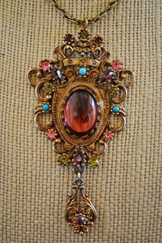 This necklace includes a large purple cabochon stone in the center along with small turquoise beads, purple crystals, pink, and green crystals.  Chain is the same matching color as the pendent.  Looks like Trifari.  Beautiful vintage necklace.  Necklace is 28.5'' long (untied.)  Pendent is just over 3'' long. Cheap Antique Medallion Necklaces, Luxury Ornate Stone Work Necklaces, Collectible Ornate Pendant Locket Necklace, Antique Purple Pendant Necklace, Ornate Purple Gemstone Necklace, Victorian Garnet Necklace, Ornate Amethyst Pendant Necklace, Punk Vintage, Medallion Necklace