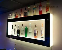 an illuminated shelf with bottles and glasses on it in front of a wall mounted mirror