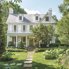 a large white house surrounded by trees and flowers in the front yard with stone steps leading up to it