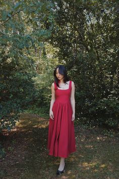 a woman in a red dress standing next to some bushes and trees with her hands on her hips