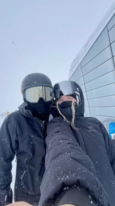 two people in jackets and helmets are standing on snow covered ground with their arms around each other