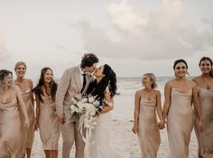 a bride and groom kissing on the beach with their bridal party in tan dresses