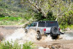 a truck is driving through some water in the mud near trees and bushes on a dirt road