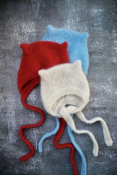 two knitted hats laying next to each other on top of a gray surface with red, white and blue yarn