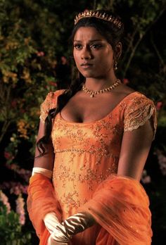 a woman in an orange dress and tiara is standing outside with flowers behind her