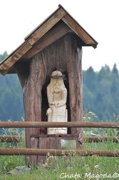 a wooden birdhouse with a statue in the center and a fence surrounding it on a grassy field