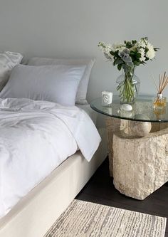 a glass table with flowers on it next to a white bed and rug in a bedroom