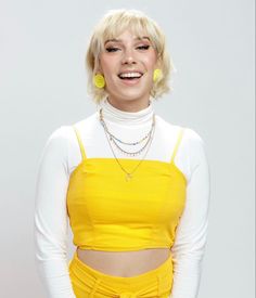 a woman with short blonde hair wearing a yellow outfit and smiling at the camera while standing in front of a white background