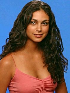 a woman with long dark hair wearing a pink top and posing for a photo in front of a blue background