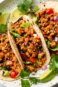 three tacos on a plate with cilantro, tomatoes and lime wedges