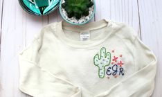 a white sweater with a cactus embroidered on it next to a potted green plant