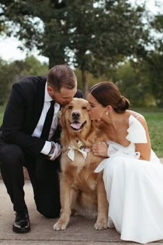 a bride and groom kissing their dog on the ground in front of some trees,