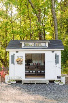 a small white shed sitting on top of a gravel road in front of some trees