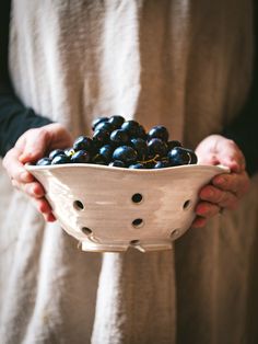 a person holding a white bowl with black berries in it and wearing an apron over their shoulders