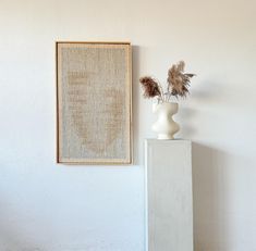 a white vase sitting on top of a cement block next to a framed art piece