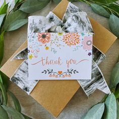 a thank you card with flowers and leaves on top of some brown envelopes next to greenery