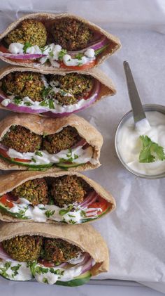 four pitas filled with meat and vegetables on top of a white napkin next to a bowl of yogurt