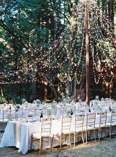 an outdoor dinner table set up with white linens and lights strung from the trees