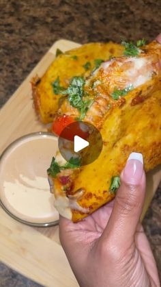 a person holding a piece of pizza on a cutting board with dipping sauce in the background