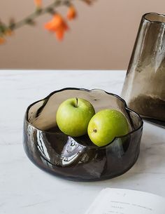 two green apples in a bowl on a table next to an open book and vase