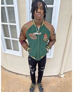 a young man with dreadlocks standing in front of a door wearing a green and brown jacket