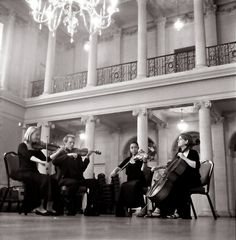 four women are playing violin in an old building with chandelier and columns on the second floor