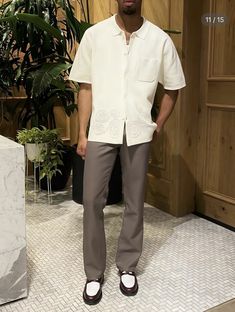 a man standing in front of a plant wearing slip ons and a white shirt