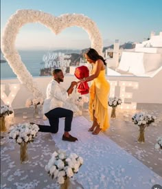 a man kneeling down next to a woman in front of a heart - shaped arch