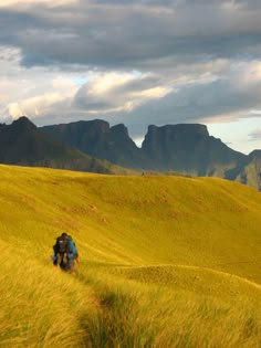 two people walking through the grass with mountains in the background and clouds in the sky