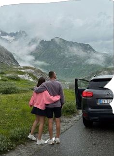 two people are standing in front of a car on the road with mountains in the background