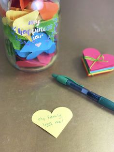 a jar filled with lots of paper hearts next to a pen and sticky notes on a table