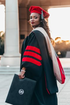 a woman in a graduation gown is holding a black briefcase and wearing a red cap