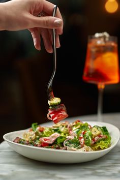 a person holding a fork full of food over a salad on a plate with a drink in the background