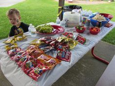 a little boy sitting at a table with food on it and bags of chips in front of him