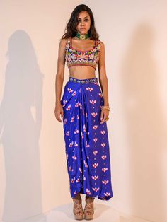 A three-piece Blue Indica Mirror Dhoti Skirt Set Paired with Bralette and Cape from the Juhi Bengani collection – Rang-Festive 2022. This gorgeous blue indica silk mirror dhoti skirt is paired with a heavy embroidered multicolored bralette blouse. The bright and vibrant colors with floral print, heavy embroidery of mirror, beads, stones, and thread sequence work enhance this satin dhoti skirt set. The indica blue dhoti skirt has a side opening at the waistline. This outfit is completed with a mu Reception Sharara With Tilla Details, Multicolor Sharara With Tilla Embroidery, Sleeveless Blue Sets For Reception, Blue Sleeveless Sets For Reception, Blue Sleeveless Set For Reception, Party Sharara With Tilla And Traditional Drape, Blue Silk Set For Navratri, Blue Fitted Pre-draped Saree For Transitional Season, Blue Palazzo Set For Reception And Festivals