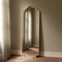 a large mirror sitting on top of a wooden floor next to a white curtained window