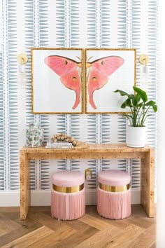 two pink stools in front of a wooden table with paintings on the wall behind it