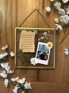 a frame with flowers and a note attached to it on a wooden table next to some white flowers