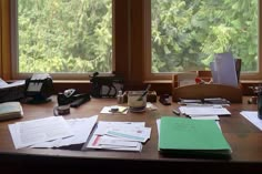 a wooden desk topped with lots of papers next to two large windows filled with trees