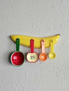 an assortment of kitchen utensils hanging on a wall next to a banana, apple, watermelon and orange