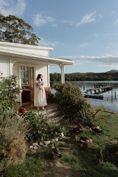 a woman standing on the porch of a house next to a body of water with boats in the background