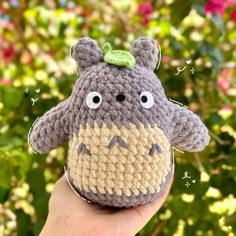 a hand holding a small crocheted stuffed animal in front of some bushes and flowers