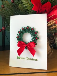 a christmas card with a red bow and green wreath on it, sitting next to a pine tree