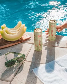 two cans of soda sitting next to a pool with bananas and sunglasses on the ground