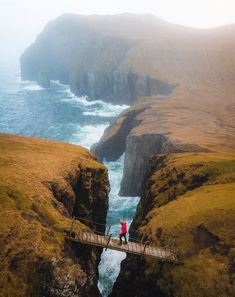 two people walking across a bridge over a body of water with cliffs in the background